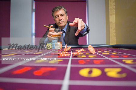 Man sitting at table holding a cigar and a glass of whiskey throwing chips