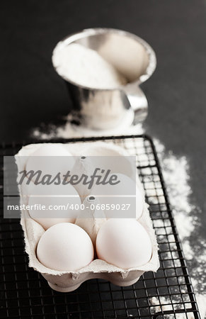 Chicken eggs in white carton on mesh tray with flour in metal measuring cup on dark background
