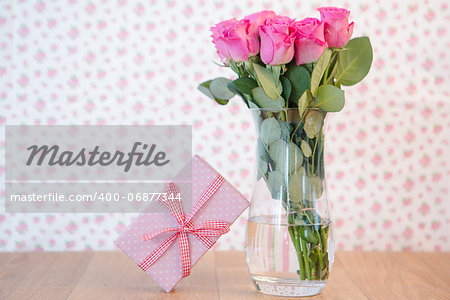 Bouquet of pink roses in vase with pink gift leaning against it on wooden table