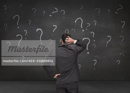 Rear view of a doubtful businessman looking at various question marks drawn on a grey wall