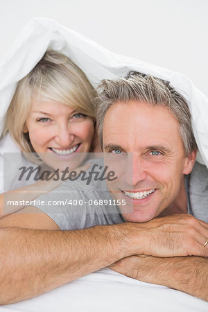 Couple smiling under the covers at the camera at home in bed