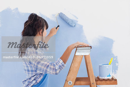 Woman using paint roller to paint wall in blue paint leaning on ladder