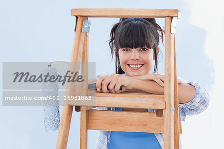 Smiling woman holding paint roller leaning on ladder looking at camera