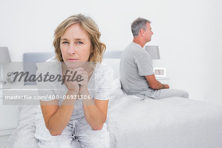 Thoughtful couple sitting on different sides of bed having a dispute with woman looking at camera in bedroom at home