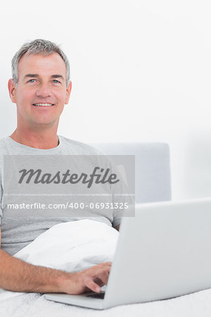 Smiling grey haired man using his laptop in bed looking at camera in bedroom at home