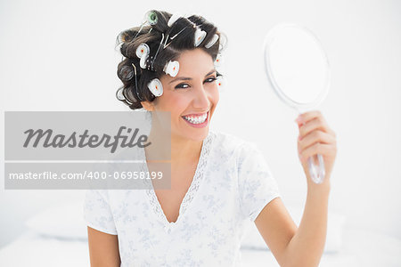 Happy brunette in hair rollers holding hand mirror smiling at camera at home in bedroom