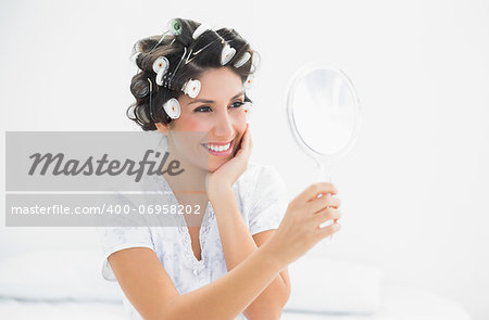 Smiling brunette in hair rollers looking in hand mirror at home in bedroom
