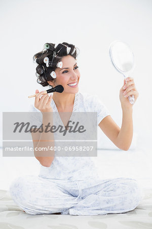 Cheerful brunette in hair rollers holding hand mirror and applying makeup at home in bedroom