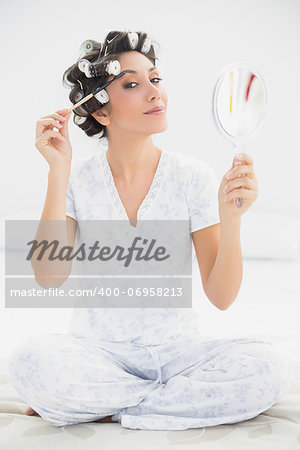 Happy brunette in hair rollers holding hand mirror and brushing her eyebrows at home in bedroom