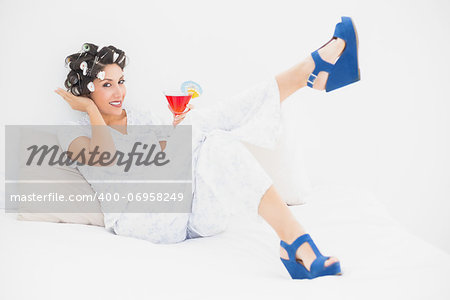 Brunette in hair rollers and wedge shoes holding a cocktail looking at camera in bedroom at home