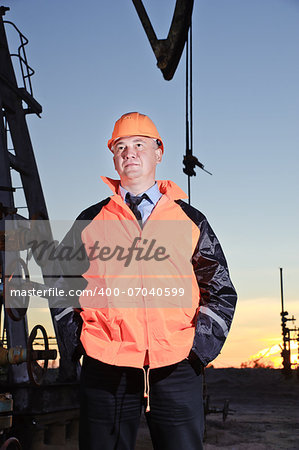 Worker in orange uniform and helmet on of background the pump jack and sunset sky. Severe. Hands in pockets.