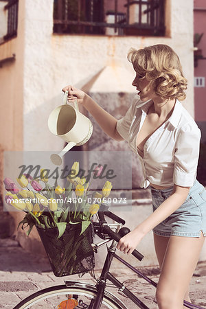 funny portrait of sexy blonde woman with denim shorts watering coloured tulips in the bicycleâ??s basket