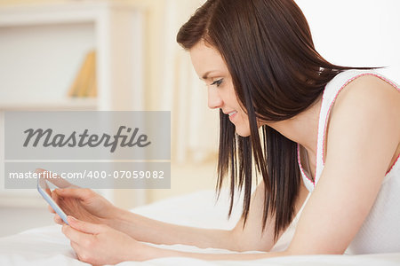 Smiling brunette using a tablet pc lying on her bed in a bedroom