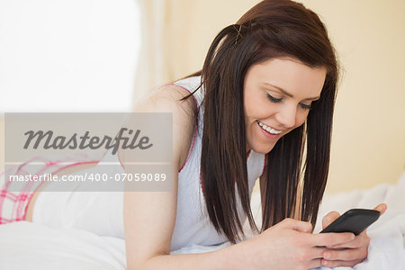 Smiling brunette using a mobile phone on a bed in a bedroom
