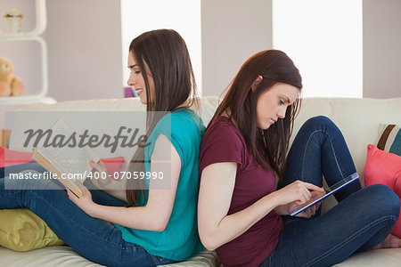 Two happy friends sitting back to back on the sofa reading book and using tablet at home in living room