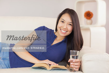 Beautiful asian girl lying on the sofa reading a novel and drinking water in living room at home