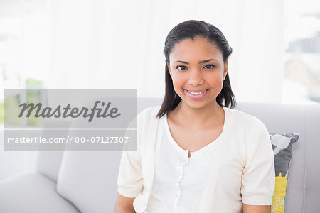 Smiling young dark haired woman in white clothes looking at camera in a living room