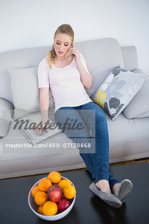 Casual pretty blonde phoning and using laptop in bright living room