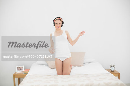 Woman playing air guitar while listening to music, kneeling on her bed and looking into the camera