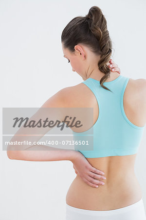 Rear view of a toned young woman with back pain standing against wall in fitness studio