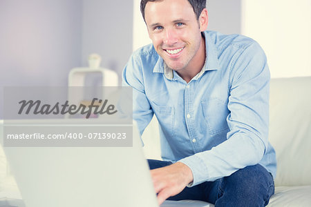 Cheerful casual man using laptop and looking at camera in bright living room