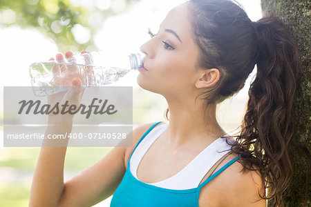 Active calm brunette drinking from a water bottle in a park on a sunny day