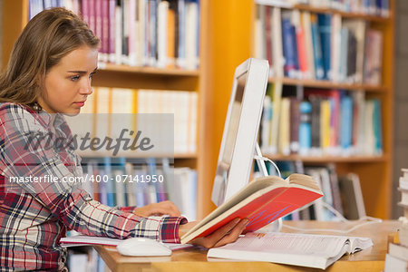 Pretty focused student reading book using computer in library