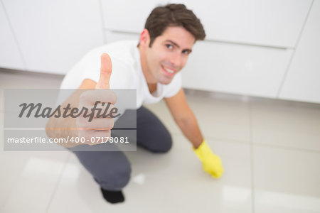 Portrait of a smiling young man cleaning the kitchen floor while gesturing thumbs up at house