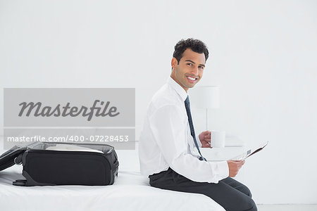 Smiling young businessman with coffee cup reading newspaper by luggage at a hotel room