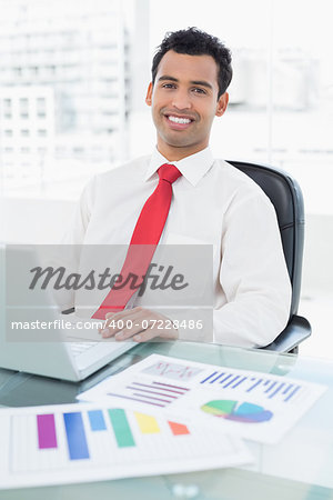 Portrait of a smiling young businessman with laptop and graphs sitting at office desk