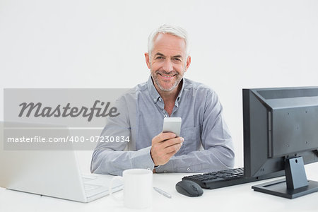 Portrait of a mature businessman with cellphone, laptop and computer at desk against white background