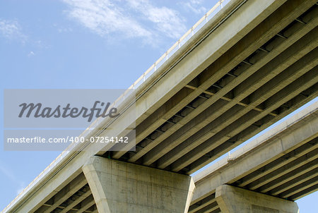 Underneath pair of large concrete highway bridges against partly cloudy blue sky.