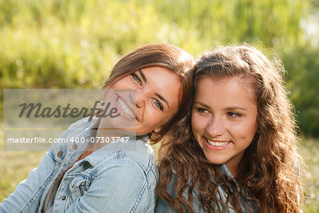 two girlfriends in jeans wear outdoors sitting back-to-back smiling