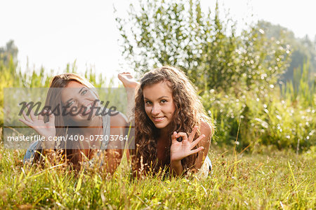 two girlfriends in T-shirts  lying down on grass showing ok sign looking at camera