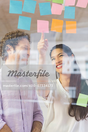 Happy businessman and businesswoman discussing over adhesive notes on glass wall in office