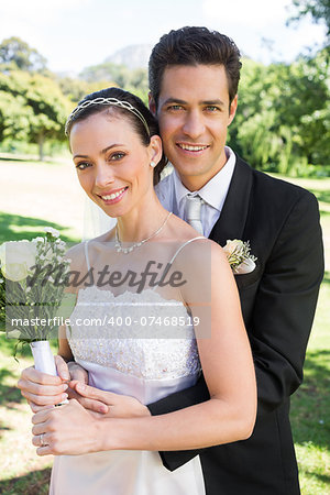 Portrait of happy groom hugging bride from behind in park