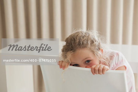 Portrait of a cute young girl hiding face behind chair in the house