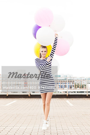 beautiful girl with ponytail hair in short black and white striped dress and white high sneakers walks forward holding bunch of multicolored balloons