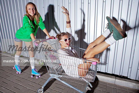 a beautiful young girl in sexy shorts and roller skates is pushing a supermarket trolley with a laughing girl. they both are projecting shadows onto the wall