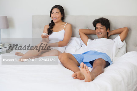 Happy couple lying on bed together watching tv at home in bedroom