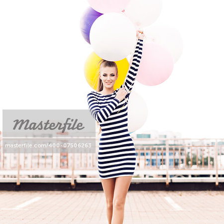 beautiful girl with ponytail hair in short black and white striped dress and white high sneakers walks forward holding bunch of multicolored balloons