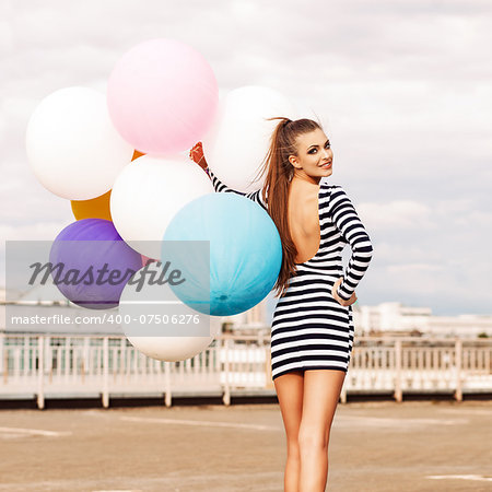 smiling girl in open-back black and white striped short dress and white high top sneakers with bunch of multicolored balloons turns back