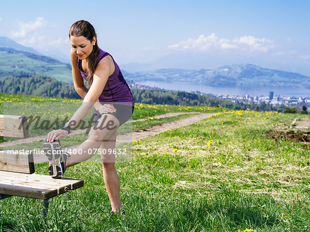 Photo of a young woman stretching her leg before she starts to run on a country path.  City and lake in the distance.