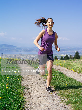 Photo of a young woman jogging and exercising on a country path.  Lake and city in the distance. Slight motion blur visible.