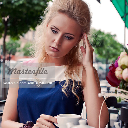 a beautiful young blond girl in summer dress at the table in pavement cafe is having a headache