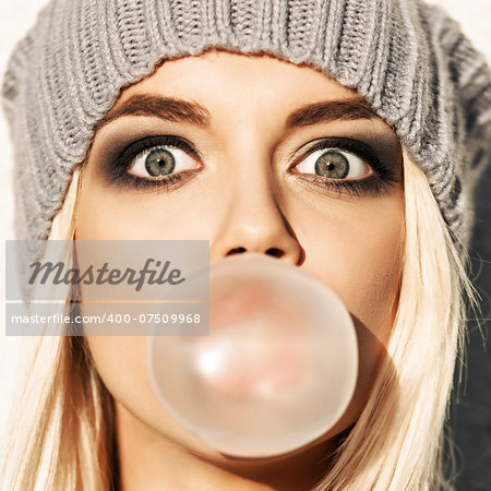 Portrait of beautiful young blond girl in beanie hat blows big bubble from bubble gum