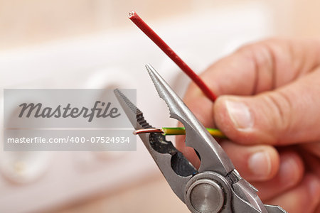 Pliers peeling copper wires - closeup on electrician hands, wall sockets in the background