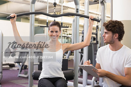 Fit brunette using weights machine for arms with trainer taking notes at the gym