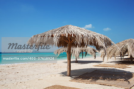 Straw beach umbrellas and mats at a tropical resort on a sandy beach overlooking an azure blue ocean on a hot summer day for a perfect getaway. Paradise island