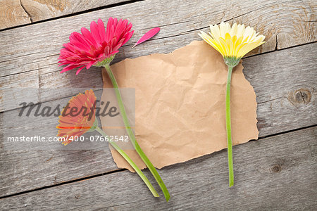 Three colorful gerbera flowers with paper for copy space on wooden table